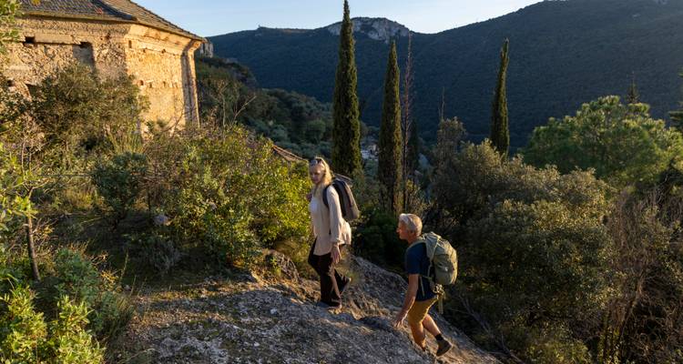 Deux randonneurs avec des sacs à dos remontant un sentier rocheux en passant devant de grands cyprès au coucher du soleil.