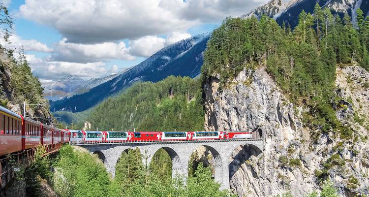 Train panoramique rouge traversant un haut viaduc de pierre au milieu d'une forêt alpine et de montagnes escarpées