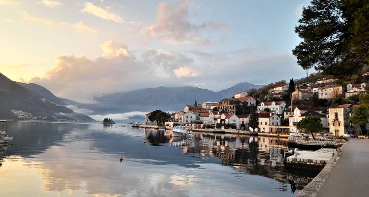 Charming stone village of Perast reflected in calm sunset waters with misty mountains behind.