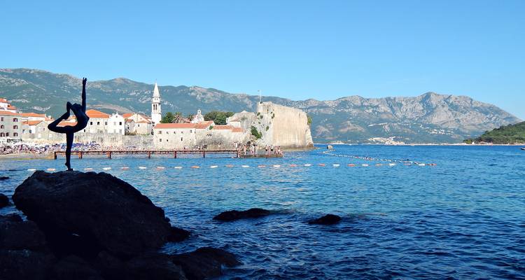 Scenic coastal view of Budva old town with the famous ballerina statue on a rock in the foreground and turquoise Adriatic waters.
