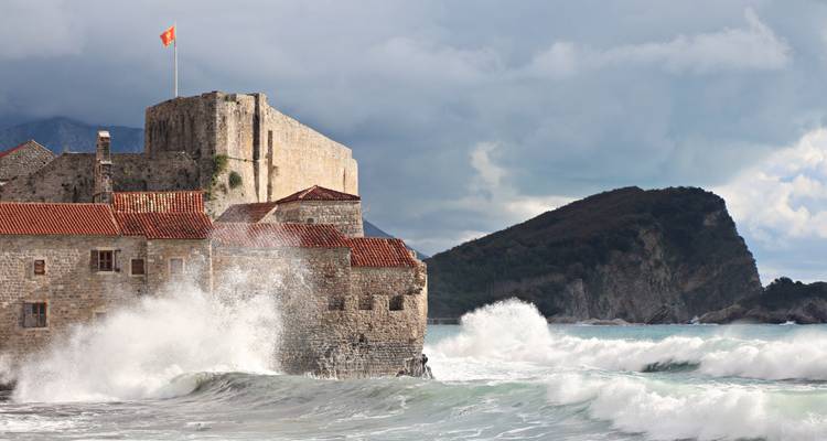 Dramatic waves crash against the medieval stone walls of Budva citadel crowned with a Montenegrin flag.