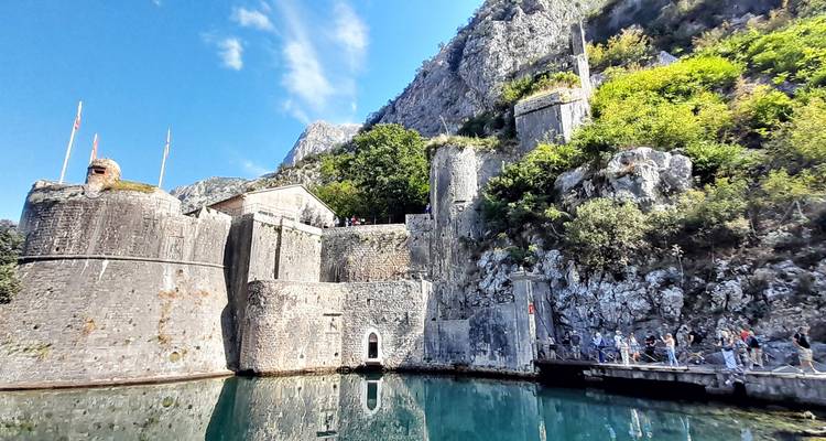 Visitors walk along a footbridge toward the stone fortifications and emerald moat of Kotor’s city walls backed by steep cliffs.