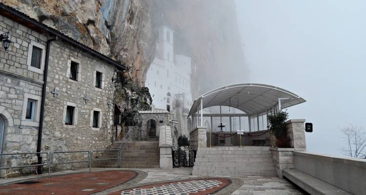The white Ostrog Monastery built into a sheer cliff face emerges through thick mountain fog.