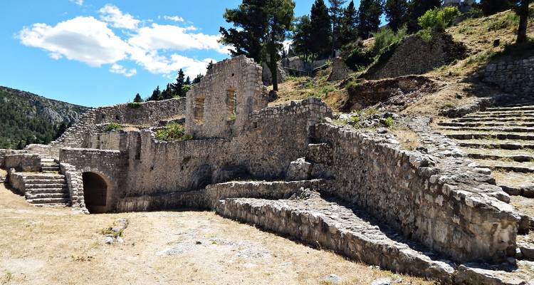 Ruinas de piedra erosionada de una antigua fortaleza en la cima de una colina yacen bajo pinos y un cielo brillante de verano.