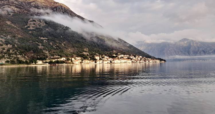 Cloud-topped mountains descend to a coastal village whose reflection ripples across calm bay waters.