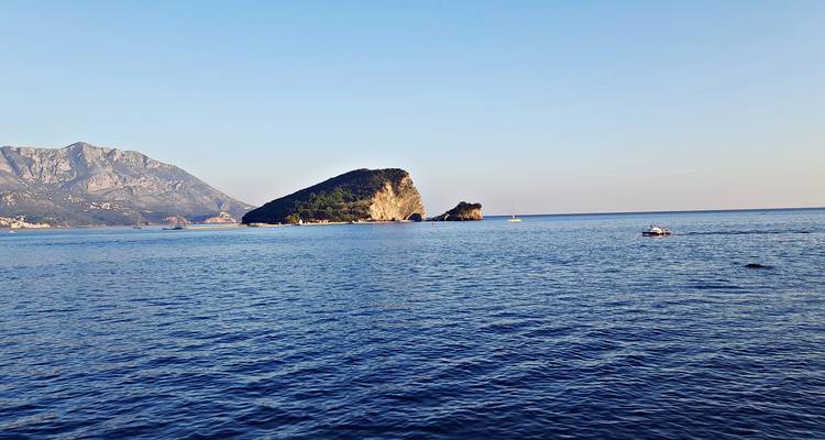 Small rocky island and sailboat on a calm Adriatic horizon with distant mountains.