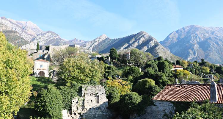 Green hillside dotted with stone ruins beneath serrated mountain peaks and blue sky.