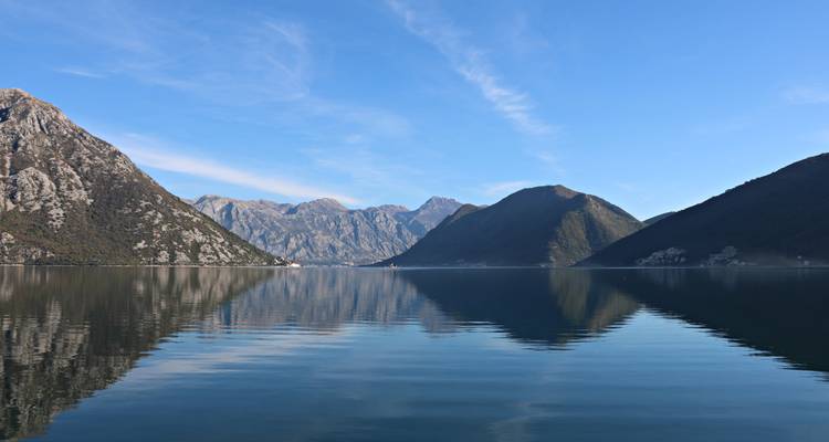 Calm bay with mirror-like reflections of steep rocky mountains under a clear blue sky.