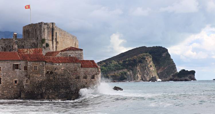 Stone fortress of an old coastal town with waves crashing against its walls and a distant rocky headland.