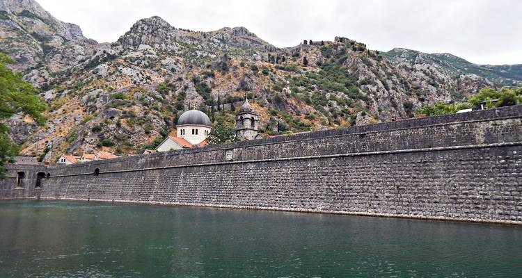Long medieval stone wall and church dome set against rocky mountains beside still water.