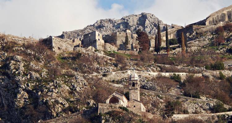 Stone ruins and winding fortification walls climbing a steep, rocky hillside beneath rugged peaks.
