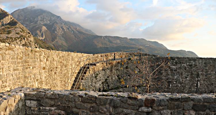 Sunlit stone walls and staircase of a hilltop fortress with dramatic mountain backdrop.
