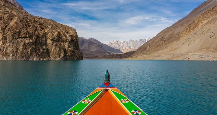 Proue de bateau en bois colorée fendant un lac turquoise flanqué de pics dénudés imposants.