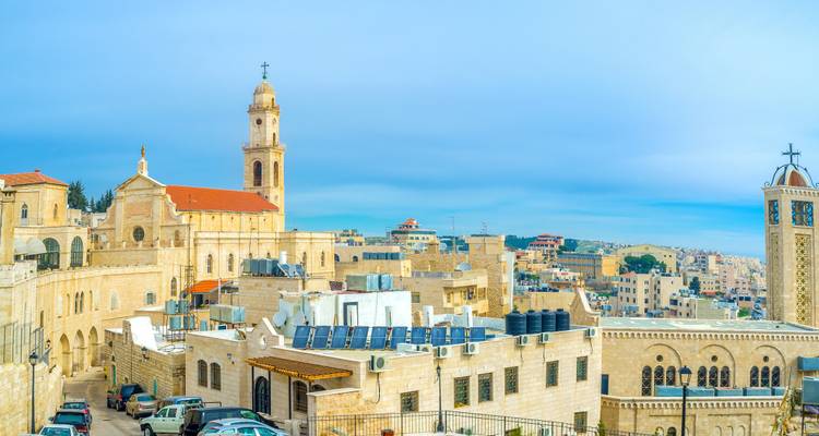 Aus Stein gebaute nahöstliche Stadtlandschaft mit braunen Gebäuden, Glockentürmen und Wassertanks unter einem ruhigen blauen Himmel.