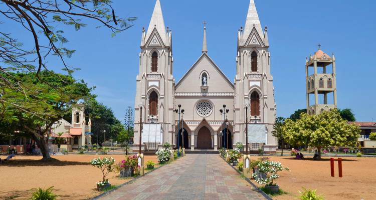 Zweiturmige Kirche im gotischen Stil, eingerahmt von einem von Bäumen gesäumten Weg und strahlend blauem Himmel auf einem sandigen Platz.