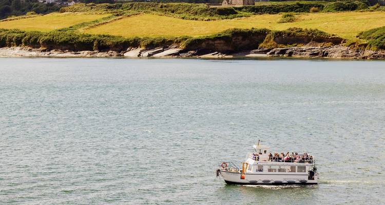Pequeño barco turístico blanco con pasajeros navega por una amplia bahía irlandesa rodeada de campos verdes.