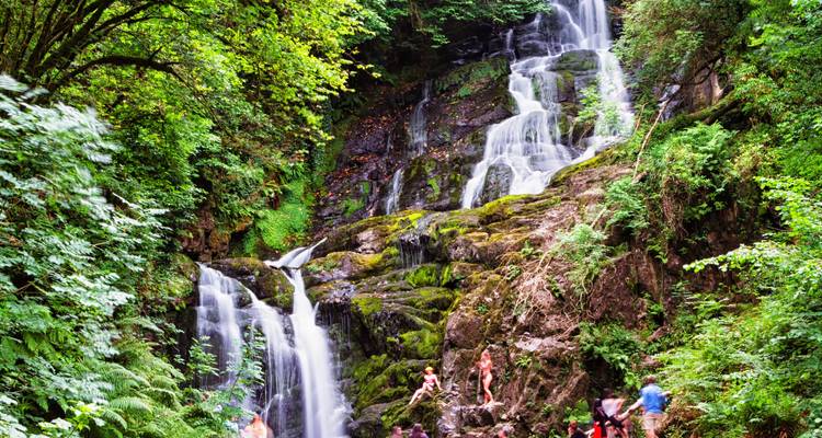 Una cascada de varios niveles se derrama a través de un exuberante bosque verde mientras los visitantes exploran las rocas de abajo.