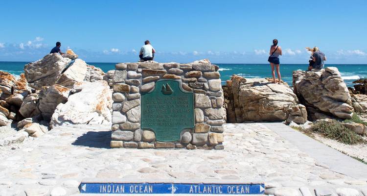 Marqueur en pierre au cap des Aiguilles montrant le point de rencontre des océans Indien et Atlantique avec des visiteurs perchés sur les rochers derrière.