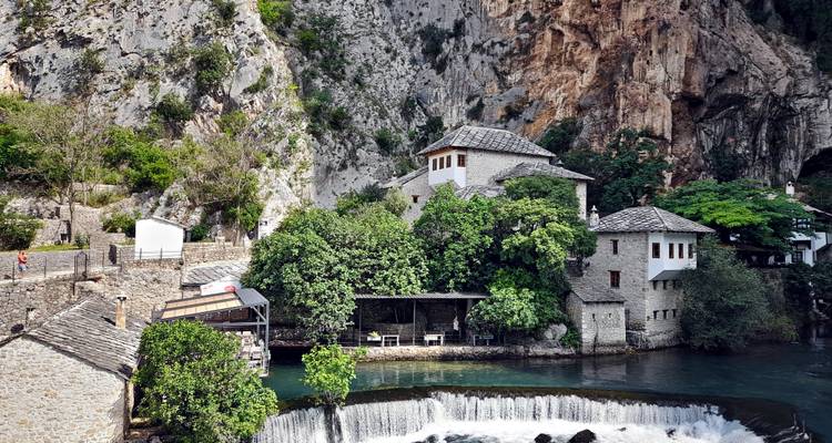Water cascades over a small weir beside Blagaj Tekke framed by lush greenery and limestone cliffs.