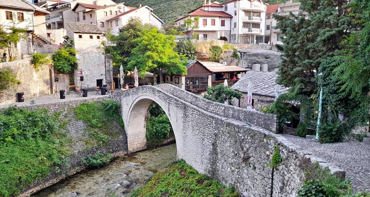 A narrow stone footbridge arches over a clear stream in a quaint Bosnian village draped with greenery.
