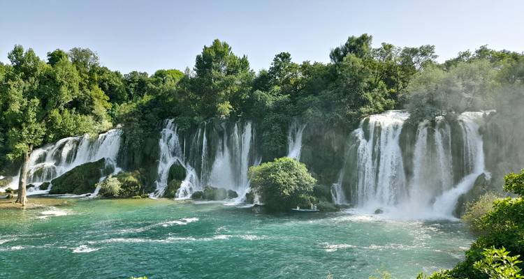 Powerful curtains of water plunge into a turquoise pool at Kravice Waterfalls surrounded by dense forest.