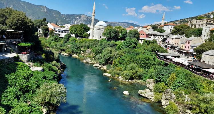A turquoise river cuts through Mostar’s old town with a mosque’s minaret rising amid stone houses and distant mountains.