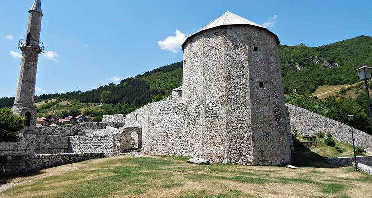 Des remparts de pierre et une tour ronde de la forteresse de Travnik s'élèvent à côté d'un minaret élancé contre le ciel bleu d'été.