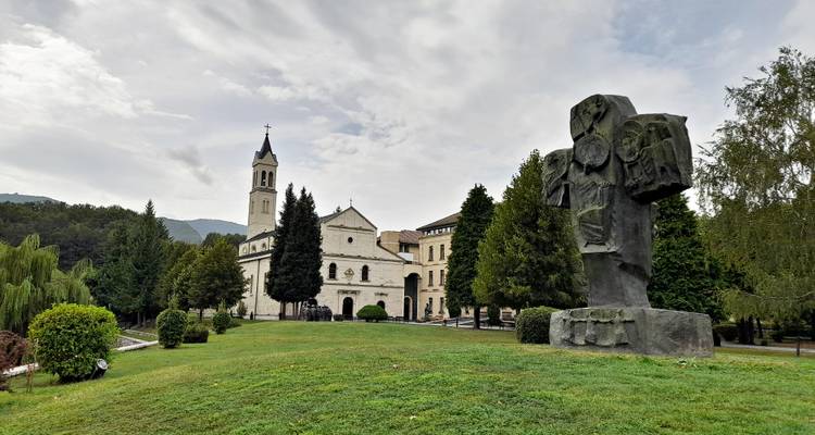 Un complexe monastique et une sculpture moderne de croix en pierre situés dans un parc paysager verdoyant avec des collines boisées en arrière-plan.