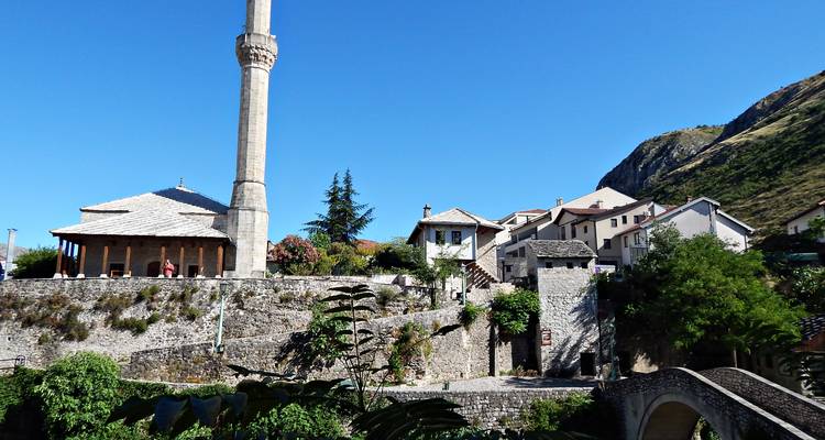 Des maisons aux murs blancs et un grand minaret se dressent sous un ciel bleu dégagé près d'un petit pont de pierre.