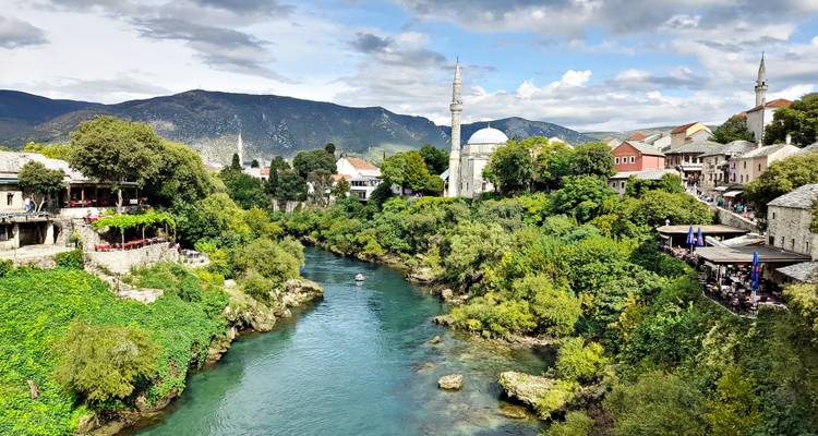 Des rives verdoyantes encadrent la Neretva turquoise qui serpente, avec des minarets et la vieille ville de Mostar au loin.