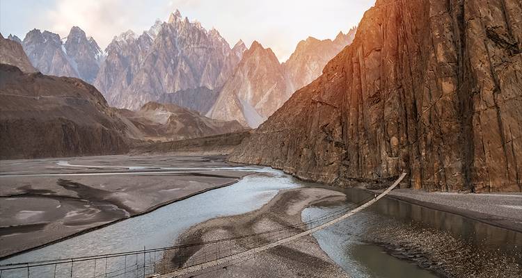 Puente colgante peatonal cruza un río glacial bajo los dramáticos Conos de Passu en la hora dorada