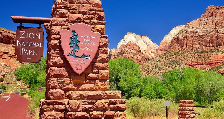 Panneau d'entrée du parc national de Zion avec des montagnes de roche rouge et un ciel bleu profond au-delà.