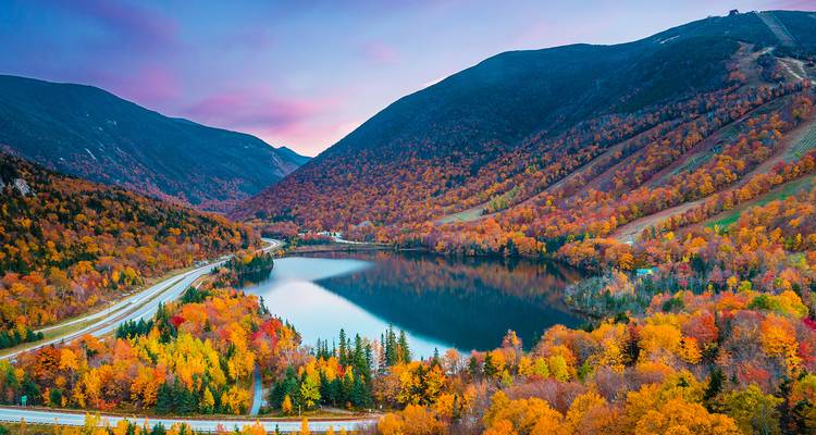 Reflektierender Bergsee umgeben von feurigem Herbstlaub und Talstraßen.