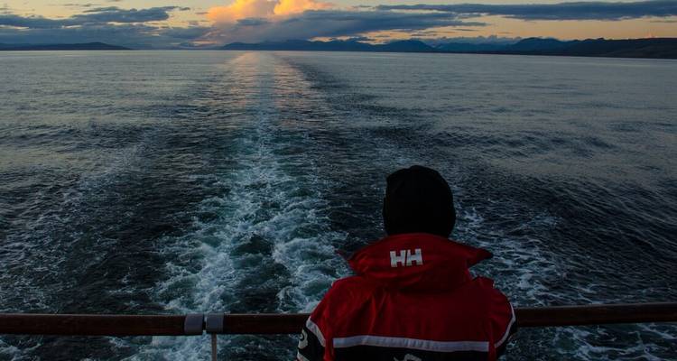Personne en veste rouge à la poupe du navire regardant le sillage se fondre dans l'horizon du coucher de soleil.