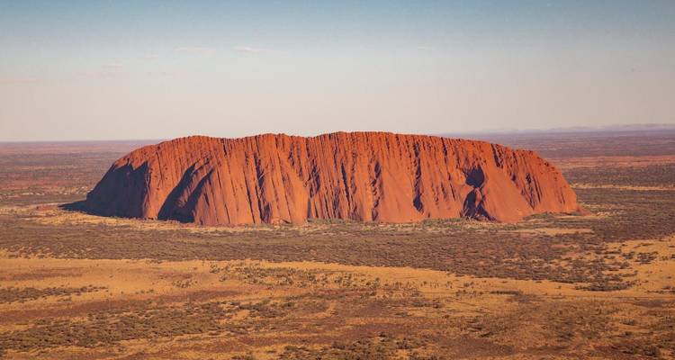 Le monolithe rouge emblématique d'Uluru s'élève des paysages désertiques plats sous un ciel bleu clair.