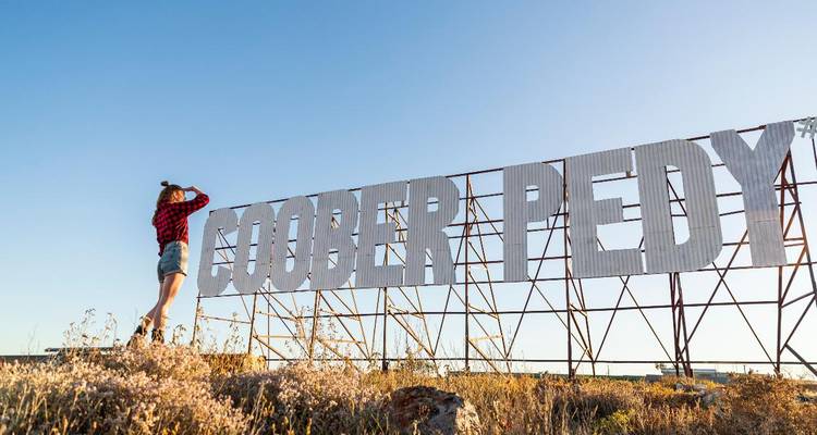 Voyageur photographiant le grand panneau métallique "COOBER PEDY" contre un ciel bleu dégagé.