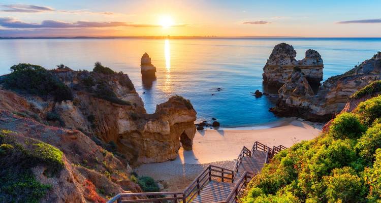 Escaleras de madera descienden a una cala aislada del Algarve mientras el sol sale sobre las tranquilas aguas del Atlántico.