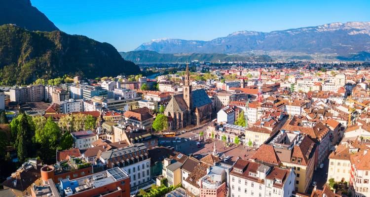 Vue aérienne du centre historique de Bolzano et de la cathédrale sur fond de montagnes alpines et de ciel bleu dégagé.