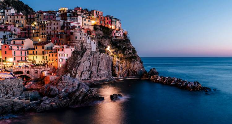 Vue crépusculaire des maisons colorées à flanc de falaise de Manarola dans les Cinque Terre illuminées au-dessus d'une mer bleue calme.