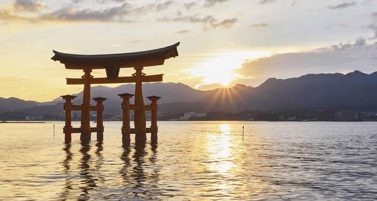Porte torii flottante emblématique du sanctuaire d'Itsukushima se découpant en silhouette contre un coucher de soleil lumineux sur des eaux calmes.