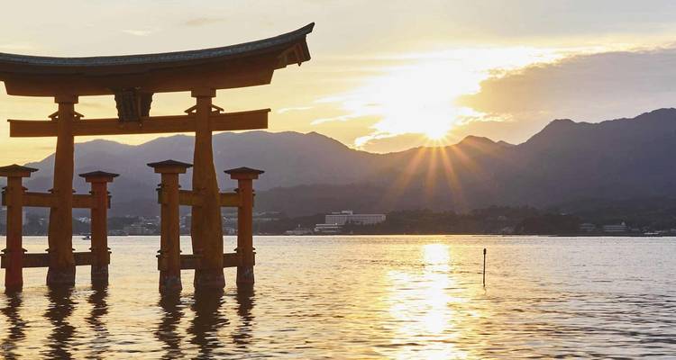 Silueta del famoso torii flotante de pie en aguas tranquilas al atardecer, con rayos de sol atravesando las crestas montañosas distantes y reflejos dorados en la superficie.