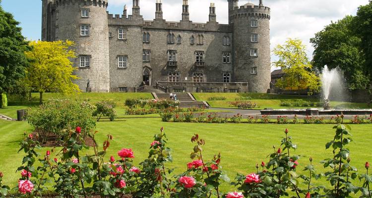 Castle with round towers behind manicured rose garden and fountain under blue sky.