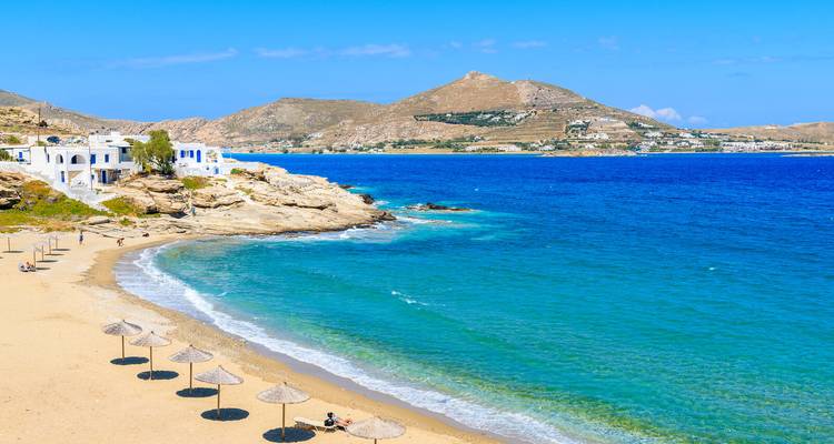 Plage de sable avec parasols en paille à côté d'eaux bleues éclatantes et de collines sur une île grecque.