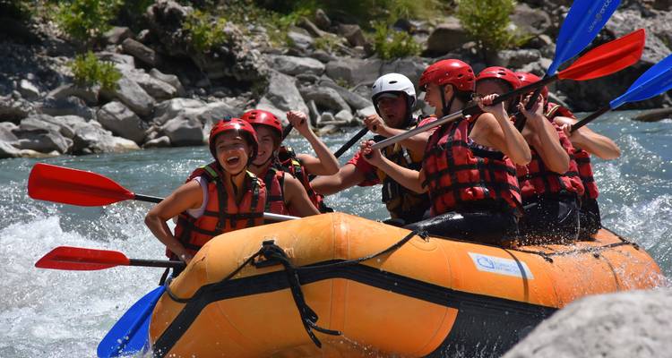 Un groupe de jeunes aventuriers faisant du rafting en eau vive dans un radeau pneumatique orange sur une rivière turquoise au courant rapide entourée de rives rocheuses.
