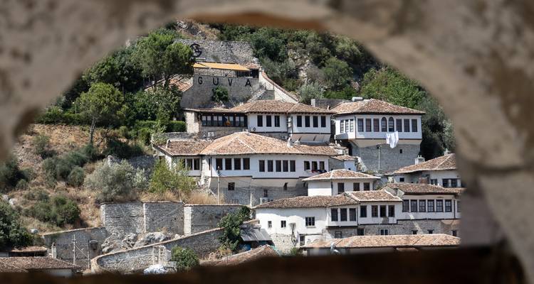 Maisons traditionnelles en pierre de style ottoman regroupées sur une colline verdoyante, encadrées par une arche de pierre brute.