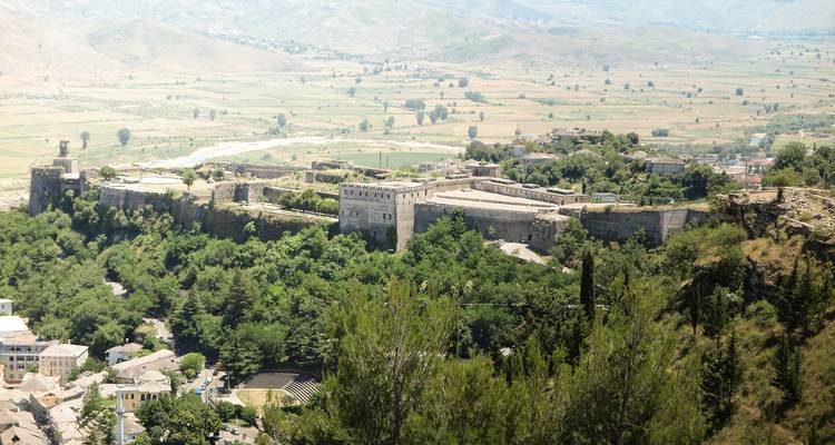 Vue en plongée d'une vaste forteresse de pierre couronnant une crête verdoyante au-dessus d'une large plaine fluviale.