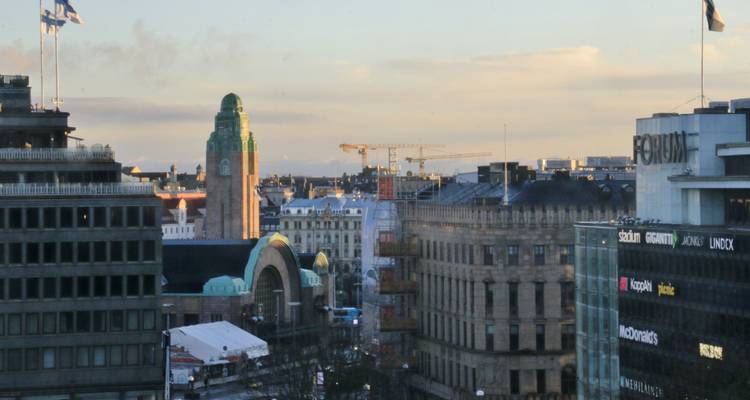 Paysage urbain du centre-ville d'Helsinki avec la tour de la gare ferroviaire au toit vert et des grues sous un ciel pastel.