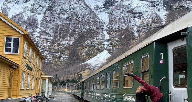 Voyageur s'étirant joyeusement à côté du wagon vert du chemin de fer de Flåm avec des montagnes enneigées qui se dressent derrière.