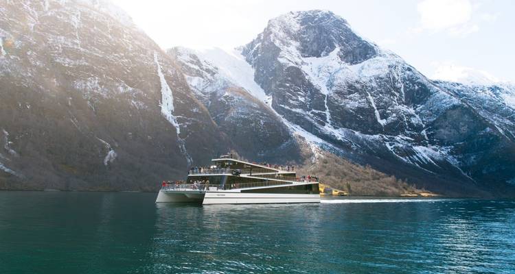 Ferry électrique moderne naviguant sur un fjord calme sous d'imposantes montagnes enneigées éclairées par une lumière oblique.