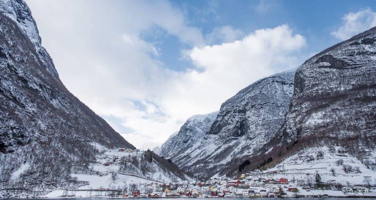 Village enneigé niché au pied de parois abruptes de fjord sous un ciel d'hiver partiellement nuageux.